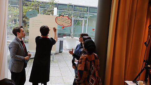 Three people stand in front of a window discussing while one of them points to a poster with colorful speech bubbles.