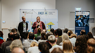 Zwei Vortragende vor sitzendem Publikum auf einer Bühne.