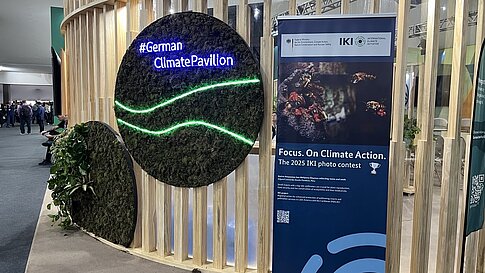 Moos-Wand mit Schriftzug "#GermanClimatePavilion" neben einem Roll-up-Banner der IKI zum Fotowettbewerb 2025