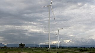 Mehrere Windkraftanlagen stehen auf einem weiten grünen Feld unter einem bewölkten Himmel. Im Hintergrund sind Berge zu sehen, während die riesigen Turbinen die erneuerbare Energieproduktion symbolisieren.