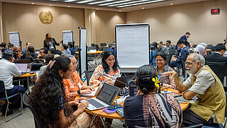 Menschen sitzen in kleinen Gruppen an runden Tischen in einem Tagungsraum und arbeiten gemeinsam an Aufgaben, während eine Frau im Hintergrund vor einem Flipchart steht.