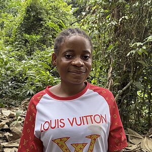Eine Frau mit kurzen, geflochtenen Haaren steht in einem Wald. Sie trägt ein weißes T-Shirt mit roten Ärmeln und dem Schriftzug 'Louis Vuitton' in goldener Farbe auf der Vorderseite. Im Hintergrund sind dichte grüne Pflanzen und Bäume zu sehen, der Boden ist mit trockenen Blättern bedeckt.