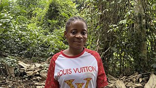 Eine Frau mit kurzen, geflochtenen Haaren steht in einem Wald. Sie trägt ein weißes T-Shirt mit roten Ärmeln und dem Schriftzug 'Louis Vuitton' in goldener Farbe auf der Vorderseite. Im Hintergrund sind dichte grüne Pflanzen und Bäume zu sehen, der Boden ist mit trockenen Blättern bedeckt.