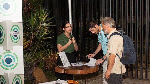 A staff member at an accreditation table talks with two arriving attendees, one of whom is filling out a form. Pens, documents, and white umbrellas rest on the table, with plants and a black fence in the background.