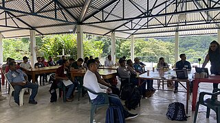 Gruppe von Menschen sitzt in einem offenen, überdachten Raum bei einer Schulung mit Tischen, Stühlen und einem Laptop auf einem Tisch