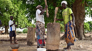 Two women stand under a tree using large wooden pestles in a hollow tree trunk while two children stand nearby watching
