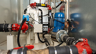 A complex system of pipes, valves, pumps, and electrical components in an industrial or technical environment. The components are connected with various cables and hoses.