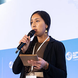 Zwei Frauen stehen auf einer Bühne bei der Global NDC Conference 2025. Eine Frau spricht ins Mikrofon, während sie ein Tablet hält; die andere Frau steht daneben mit einem Tablet in den Händen. Hinter ihnen ist eine blaue Wand mit dem Logo der Konferenz und zwei weißen Sesseln sichtbar.