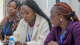 Drei Frauen sitzen bei einer Veranstaltung auf dem Podium und sprechen zum Publikum.