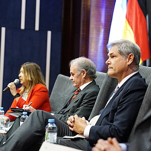 A group of four people, including three men and one woman, are seated at a podium participating in an official event. In the background, two flags are visible.