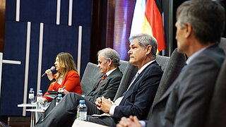 A group of four people, including three men and one woman, are seated at a podium participating in an official event. In the background, two flags are visible.