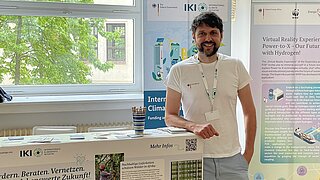 Mann mit Bart und T-Shirt steht lächelnd vor einem Messestand mit Informationsplakaten und Flyern in einem hellen Raum mit Fenster und grünen Bäumen draußen