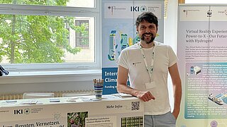 Bearded man in a t-shirt smiling while standing in front of an exhibition booth with informational posters and flyers in a bright room with a window showing green trees outside