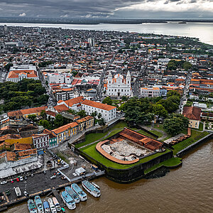 Aerial view of the historic downtown of Belém, Brazil, showcasing the cityscape and harbor. In the foreground is the Forte do Castelo (Fort) and the harbor area with boats. The streets are lined with colonial buildings, and in the background, modern skyscrapers are visible.