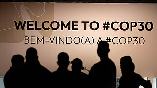 In the foreground, dark silhouettes of six people can be seen. In the background, the words "Welcome to #COP30" in English and "Bem-vindo(a) #COP30" in Portuguese can be seen.