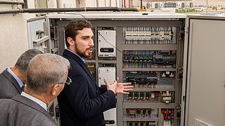 Männer stehen vor einem offenen Schaltschrank mit sichtbaren Kabeln und elektronischen Bauteilen