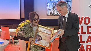 A woman receives a certificate and a prize from a man at an event while holding flowers.