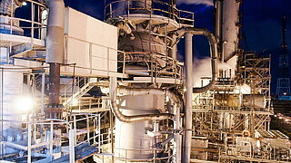 Eine komplexe industrielle Struktur bei Nacht, in der verschiedene Rohre und Metallplattformen sichtbar sind. Der Dampf, der aus der Fabrik aufsteigt, ist im Kontrast zum dunklen Himmel. Das Bild zeigt die detaillierte Architektur einer Düngerproduktionsanlage.