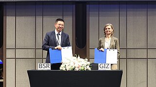 Zwei Personen stehen hinter einem Tisch mit Blumenarrangement und halten jeweils eine blaue Mappe, auf dem Tisch stehen Schilder mit den Aufschriften 'BSR' und 'GIZ'.