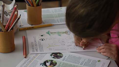 A child with sits at a table, coloring on a piece of paper. In the background, additional papers can be seen, some containing information about the environment and climate protection.
