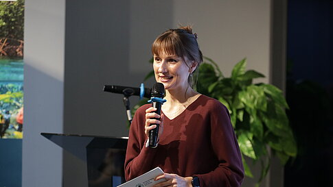 Woman holding microphone and note card speaking in front of green plants