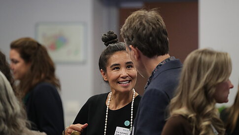 Woman with bun and pearl necklace talking to a man, surrounded by other people indoors