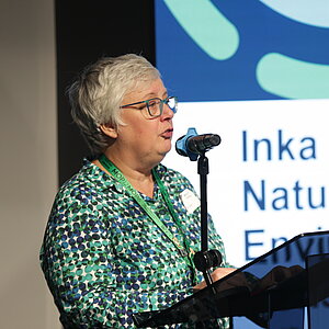Woman with short gray hair and glasses speaking at a microphone in front of a screen with text.
