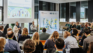 Speaker presenting in front of an audience with a large screen behind them.