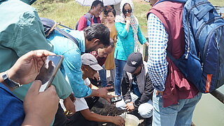 A group of people, including men and women, gather outdoors on a sunny day around a silver bucket of water. Two men are kneeling and measuring water quality with a device, while others watch or film with a smartphone. In the background, a woman holds a parasol.