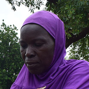 Woman wearing a blue headscarf and patterned skirt holds a metal bowl at a round mud house with a thatched roof where a man in a blue shirt and red scarf passes branches through the window