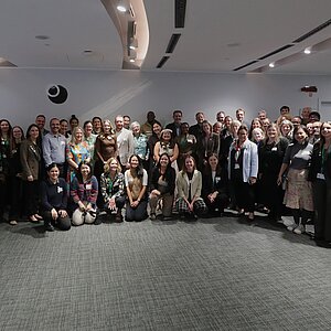 Große Gruppe von Menschen in einem Konferenzraum, einige sitzen, andere stehen, alle blicken zur Kamera