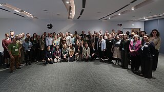 Large group of people in a conference room, some sitting and others standing, all facing the camera