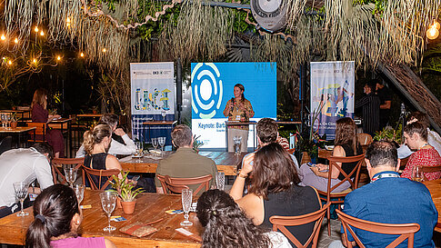 A speaker stands on stage speaking into a microphone. Behind her are banners of the International Climate Initiative. In front of her the audience sits at wooden tables with glasses on them. Green plants are visible at the edge of the image.