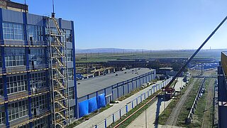 Ein Blick auf ein Industriegebäude mit einem Baukran, der gerade dabei ist, Materialien zu bewegen. Die Umgebung zeigt eine weitläufige Industriezone.