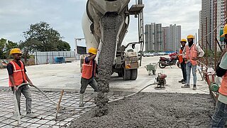 Bauarbeiter mit Schutzhelmen verteilen Beton, der aus einem Betonmischer auf ein Stahlgitter fließt, im Hintergrund Hochhäuser und Motorräder