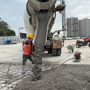 Bauarbeiter mit Schutzhelmen verteilen Beton, der aus einem Betonmischer auf ein Stahlgitter fließt, im Hintergrund Hochhäuser und Motorräder