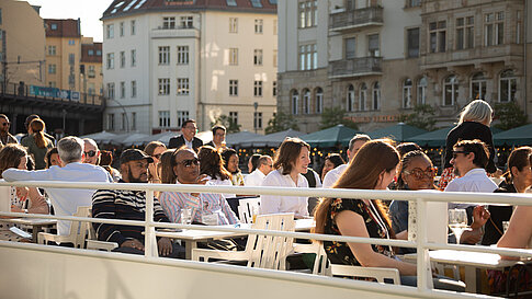Menschen sitzen im Freien auf einem Boot bei Sonnenschein. Sie unterhalten sich in kleinen Gruppen, während im Hintergrund Altbauten und ein Bahnhof zu sehen sind.