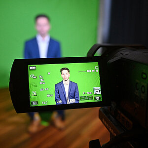 Durch den Bildschirm einer Videokamera sieht man einen Mann auf einem Stuhl vor einem grünen Hintergrund sitzen.t