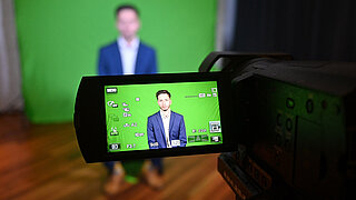 Durch den Bildschirm einer Videokamera sieht man einen Mann auf einem Stuhl vor einem grünen Hintergrund sitzen.