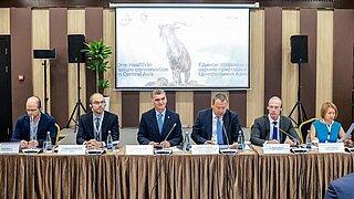 6 people are sitting on a podium. Behind them is a screen showing a presentation with an ibex. 