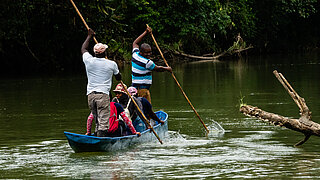 Eine Gruppe von Menschen fährt in einem schmalen blauen Kanu über einen ruhigen Fluss, umgeben von dichter, grüner Vegetation. Zwei Männer stehen und stoßen das Boot mit langen Holzstangen vorwärts, während andere Personen sitzen und sich umschauen. Auf der Wasseroberfläche schwimmt ein großer Baumstamm.