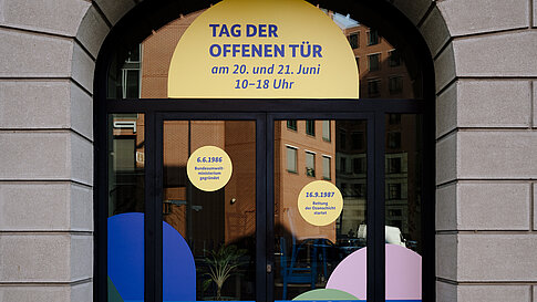 Eingangstür mit Bogen und mehreren runden Aufklebern sowie Texten über einem Schild mit der Aufschrift 'TAG DER OFFENEN TÜR'