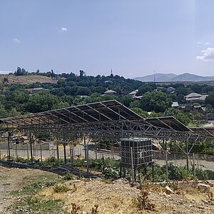 Freistehende Agri-Photovoltaikanlage mit Solarmodulen auf Metallgestell über landwirtschaftlicher Fläche in ländlicher Umgebung Armeniens mit Hügeln und Häusern im Hintergrund