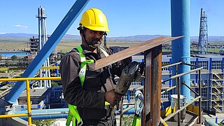 Ein Techniker arbeitet in luftiger Höhe auf einer Plattform und bearbeitet ein Metallstück mit einem Werkzeug. Im Hintergrund sind Teile des Fabrikgeländes zu sehen.