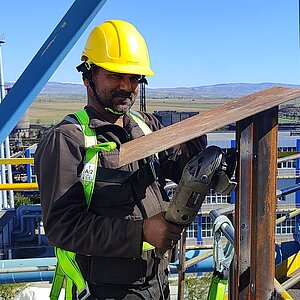 Ein Techniker arbeitet in luftiger Höhe auf einer Plattform und bearbeitet ein Metallstück mit einem Werkzeug. Im Hintergrund sind Teile des Fabrikgeländes zu sehen.