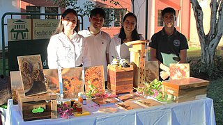Vier junge Menschen stehen hinter einem Ausstellungstisch mit Holzobjekten, Fotos von Bienen und kleinen Gläsern, vor einem Universitätsgebäude.