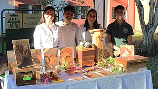 Four young people stand behind a display table with wooden objects, bee photos, and small jars, in front of a university building.