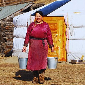 A smiling person in traditional clothing stands in front of a yurt holding two buckets in their hands.