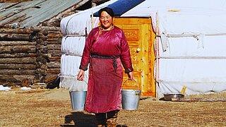 A smiling person in traditional clothing stands in front of a yurt holding two buckets in their hands.