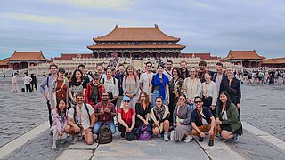 Gruppe von jungen Erwachsenen vor einem großen historischen Gebäude mit traditionellem chinesischem Dach in einer offenen Steinplatzumgebung